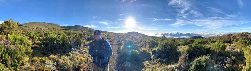 Mark Acre on his way up Kilimanjaro, standing in a landscape of grassy scrubland with a saturated blue sky in the background.