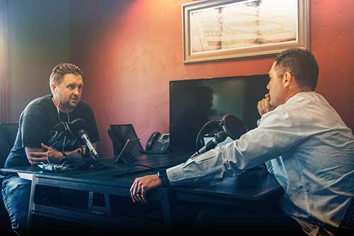 Photo of two men sitting on opposite sides of a desk. Each have a microphone and they're conducting an interview.